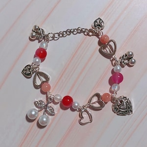 May include: A silver-toned bracelet featuring a variety of charms and beads. The bracelet includes heart-shaped charms, cherry charms, and a mix of pink, red, and white beads. The bracelet is designed with a chain and lobster clasp.