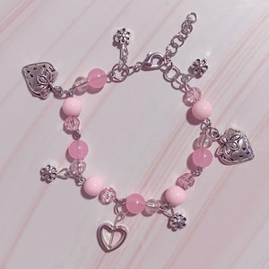 Op de afbeelding: Een roze en zilveren armband met bedels in de vorm van een aardbei, bloem en hart. De armband is gemaakt met roze kralen en een zilveren ketting.