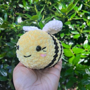 Puede incluir: Un juguete de peluche con forma de abeja hecho de hilo suave amarillo y negro con textura esponjosa. La abeja tiene dos alas blancas, pequeños ojos de plástico negro, una sonrisa negra bordada y dos pequeños círculos rosas en las mejillas.