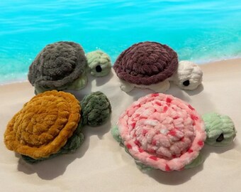 May include: Four plush crocheted turtle toys in different colors: green, brown, pink, and orange. The turtles are lying on a white sandy surface.