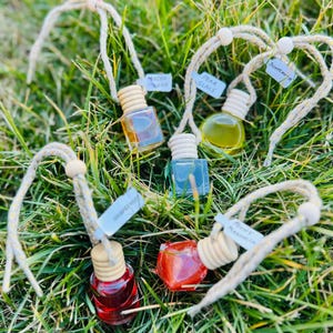 May include: Five small glass bottles filled with colorful liquid, each suspended by a twine cord. The bottles are labeled with scent names: Mucho Mango, Pear Glace, Sunflower, Grapefruit, and Ruty Magnolia. The bottles are set in green grass.