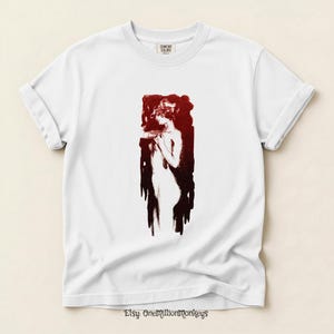 May include: White Comfort Colors t-shirt featuring a red and black illustration of a woman. The artwork is a stylized depiction of a figure with flowing lines and a dark background. The Etsy shop name "OneMillionMonkeys" is printed at the bottom.