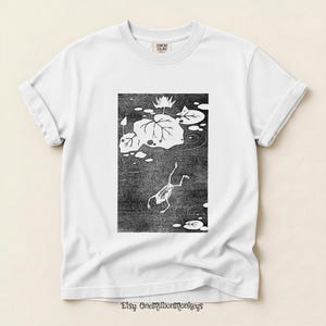 May include: White t-shirt with a black and white illustration of a frog in a pond with lily pads. The shirt has a crew neck and short sleeves. The text "Comfort Colors" is on the upper right. The Etsy shop name "OneMillionMonkeys" is at the bottom.