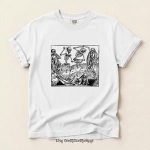 May include: White t-shirt featuring a black and white illustration of dancing skeletons. The graphic depicts several skeletons in various poses, with some appearing to dance. The shirt has a crew neck and short sleeves.