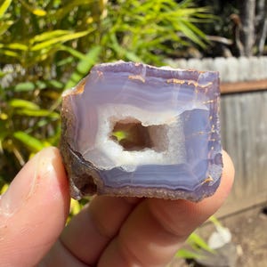 May include: A raw agate geode specimen featuring concentric bands of translucent lavender and light purple hues surrounding a central hollow cavity lined with small, sparkling white crystalline structures and rough, earthy brown outer edges.