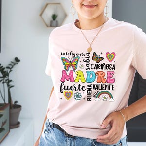 May include: Pink t-shirt with a colorful graphic design that says "MADRE" with the words "inteligente", "sabia", "cari&ntilde;osa", "fuerte", "valiente", "buena", and "bella" surrounding the word "MADRE". The graphic also includes butterflies, hearts, flowers, and a rainbow.