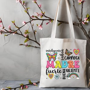 May include: White canvas tote bag with a colorful design featuring the word "MADRE" surrounded by butterflies, hearts, and flowers. The text around the word "MADRE" reads "inteligente, sabia, cari&ntilde;osa, fuerte, valiente, buena, bella".