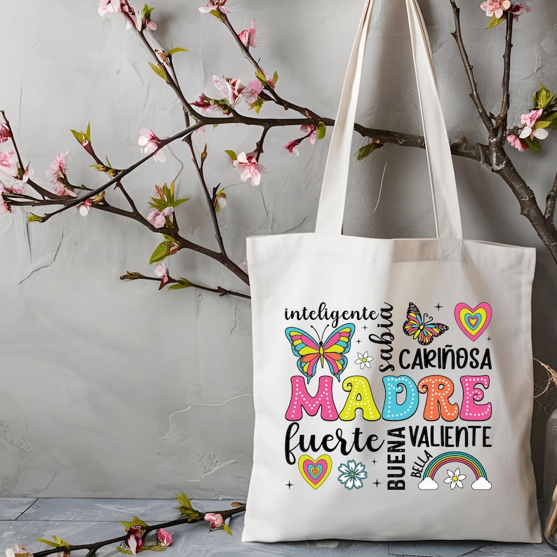 May include: White canvas tote bag with a colorful design featuring the word "MADRE" surrounded by butterflies, hearts, and flowers. The text around the word "MADRE" reads "inteligente, sabia, cari&ntilde;osa, fuerte, valiente, buena, bella".