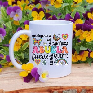 May include: White ceramic mug with a colorful design featuring the word "Abuela" surrounded by words like "inteligente", "sabia", "cari&ntilde;osa", "fuerte", "valiente", "buena", and "bella". The design also includes butterflies, hearts, and a rainbow.