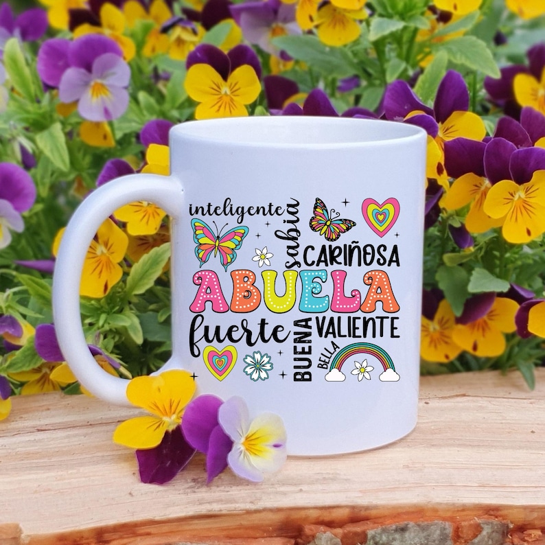 May include: White ceramic mug with a colorful design featuring the word "Abuela" surrounded by words like "inteligente", "sabia", "cari&ntilde;osa", "fuerte", "valiente", "buena", and "bella". The design also includes butterflies, hearts, and a rainbow.