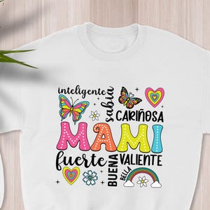 May include: White sweatshirt with a colorful graphic design that says "Mami" surrounded by words in Spanish, including "inteligente", "sabia", "cari&ntilde;osa", "fuerte", "valiente", "buena", and "bella". The design also includes butterflies, hearts, flowers, and a rainbow.