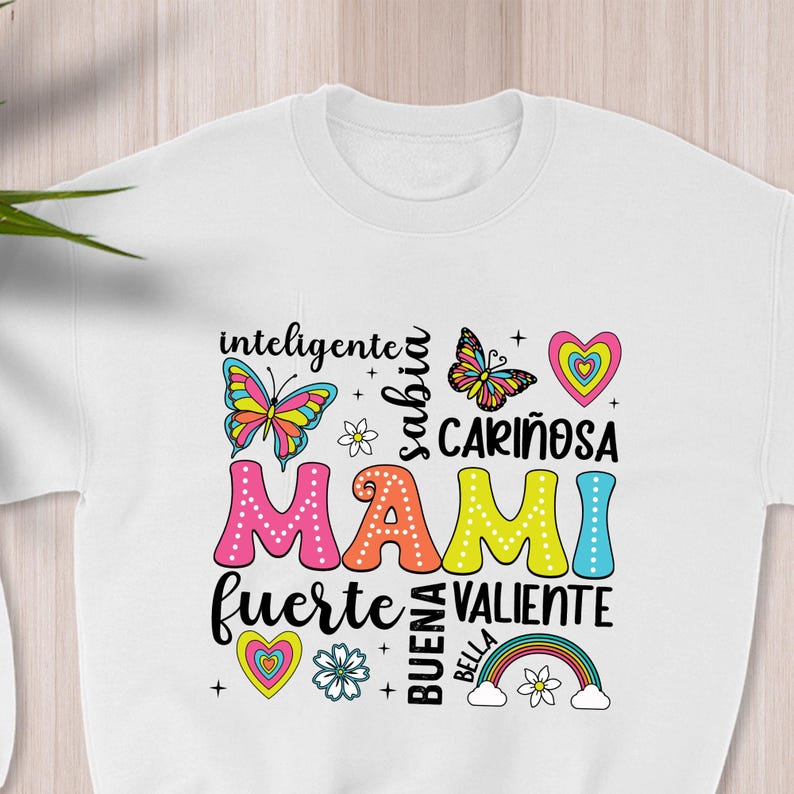 May include: White sweatshirt with a colorful graphic design that says "Mami" surrounded by words in Spanish, including "inteligente", "sabia", "cari&ntilde;osa", "fuerte", "valiente", "buena", and "bella". The design also includes butterflies, hearts, flowers, and a rainbow.