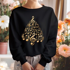 以下が含まれることがあります： 黒いスウェットシャツ。金色の音符のクリスマスツリーのデザインが特徴です。デザインには、ト音記号、音符、その他の音楽記号が含まれています。スウェットシャツはクルーネックです。
