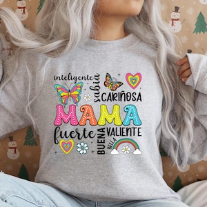 May include: Gray sweatshirt with a colorful graphic design that says "Mama" surrounded by words in Spanish, including "inteligente", "sabia", "cari&ntilde;osa", "fuerte", "valiente", "buena", and "bella". The graphic also includes butterflies, hearts, and a rainbow.