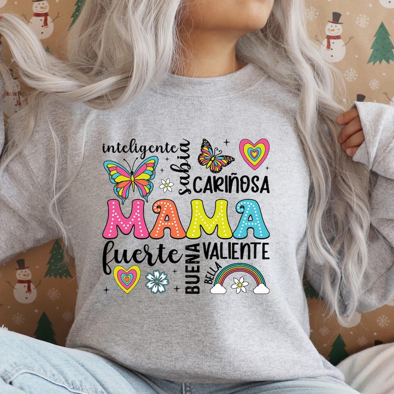 May include: Gray sweatshirt with a colorful graphic design that says "Mama" surrounded by words in Spanish, including "inteligente", "sabia", "cari&ntilde;osa", "fuerte", "valiente", "buena", and "bella". The graphic also includes butterflies, hearts, and a rainbow.