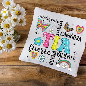 May include: White pillow with a colorful design featuring the word "TIA" surrounded by flowers, butterflies, and the words "inteligente", "sabia", "cari&ntilde;osa", "fuerte", "buena", "bella", and "valiente".