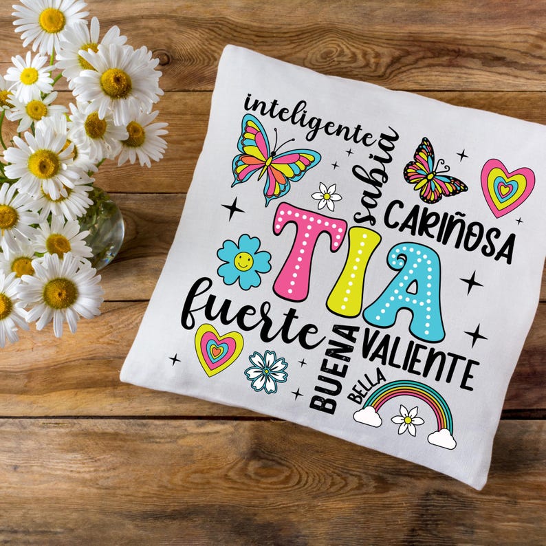 May include: White pillow with a colorful design featuring the word "TIA" surrounded by flowers, butterflies, and the words "inteligente", "sabia", "cari&ntilde;osa", "fuerte", "buena", "bella", and "valiente".