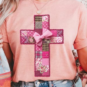 Może przedstawiać: Brzoskwiniowa koszulka z patchworkowym wzorem krzyża. Krzyż wykonany jest z różnych różowych tkanin, w tym w kropki i kwiaty, i ozdobiony różową kokardą i kwiatem. Krzyż jest obrysowany ciemniejszym różowym obramowaniem.