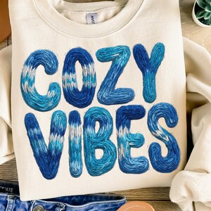 May include: Cream-colored sweatshirt with the words "COZY VIBES" embroidered in shades of blue. The letters have a textured, knitted appearance. The sweatshirt is folded, with a pair of jeans and a hat visible in the background.