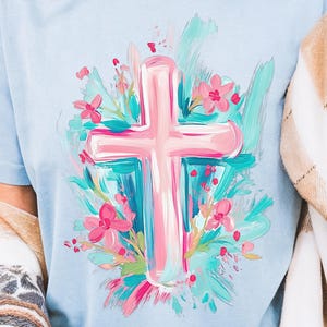 May include: A light blue t-shirt with a pink cross surrounded by blue and pink flowers. The cross and flowers are painted in a brushstroke style.