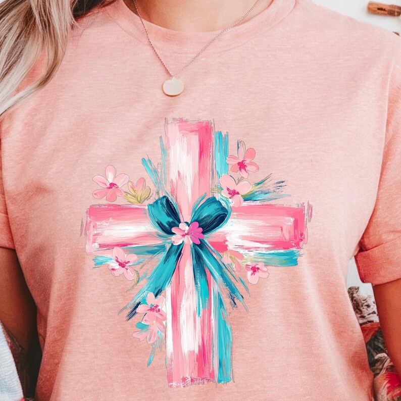 Puede incluir: Una camiseta rosa con una cruz blanca pintada con un lazo azul y flores rosas alrededor.