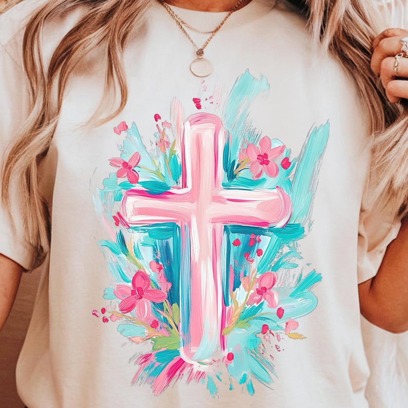 May include: A pink cross with a white outline is surrounded by blue, green, and pink brushstrokes and pink flowers. The design is on a white t-shirt.