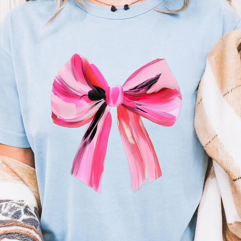 Puede incluir: Una camiseta azul claro con un gran lazo rosa y negro pintado en la parte delantera.