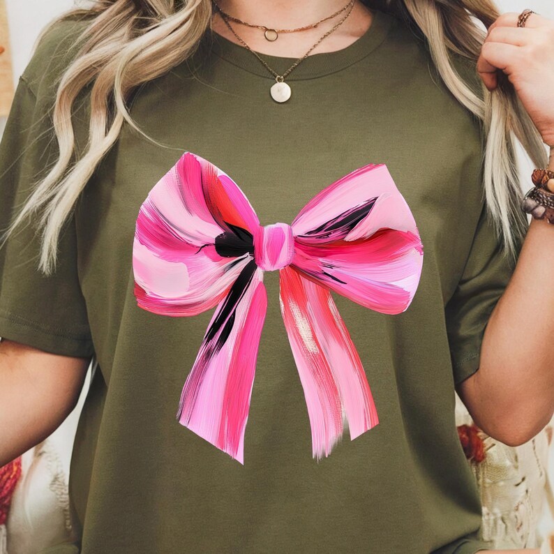 Puede incluir: Una camiseta verde con un gran lazo rosa pintado en la parte delantera. El lazo tiene un contorno negro y est&aacute; pintado con pinceladas rosas, rojas y blancas.