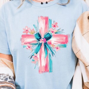 Puede incluir: Una camiseta azul claro con un dise&ntilde;o de cruz pintada en rosa y azul. La cruz est&aacute; decorada con un lazo azul y flores rosas.