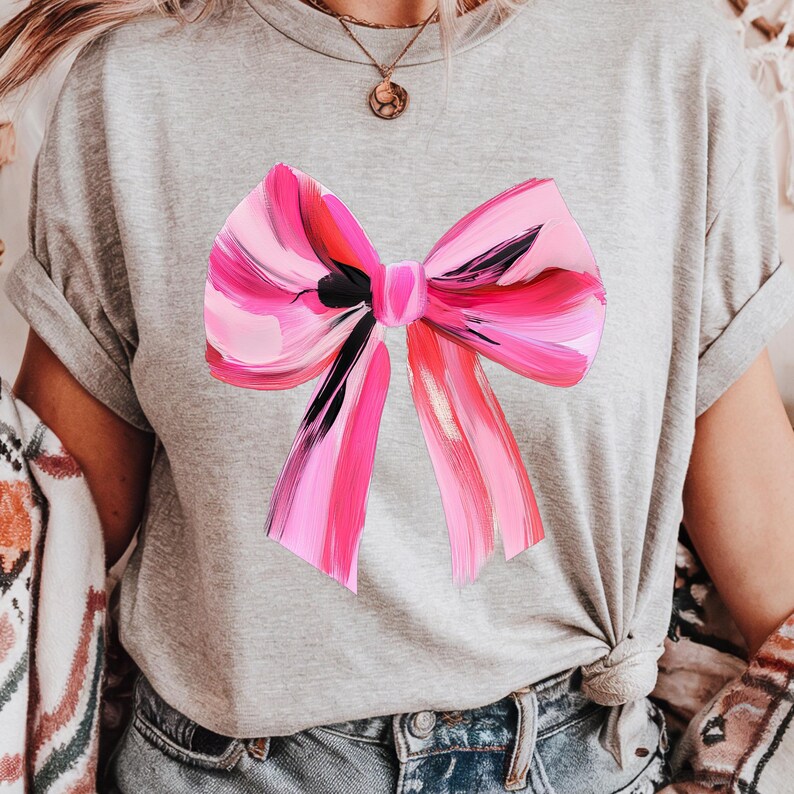 Puede incluir: Una camiseta gris con un lazo rosa y negro pintado en la parte delantera.