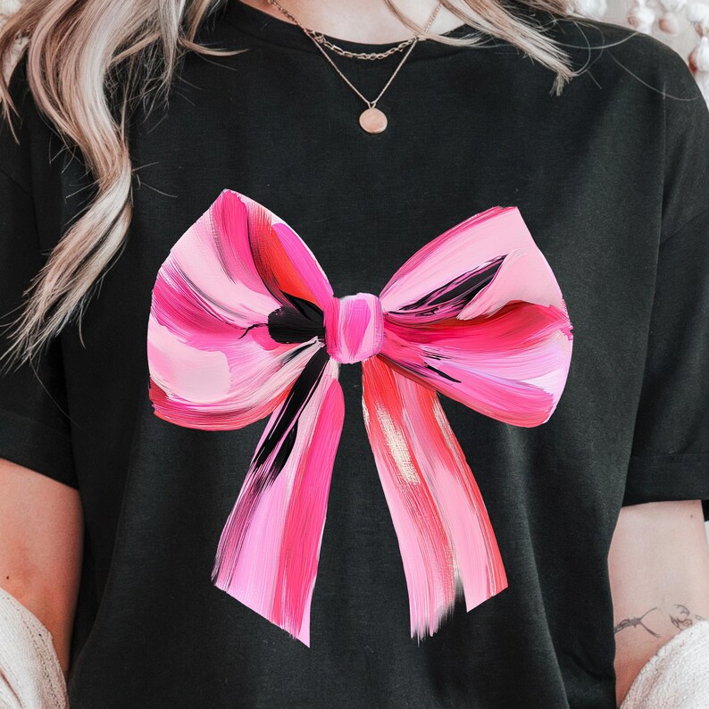Puede incluir: Una camiseta negra con un lazo rosa y negro pintado en la parte delantera. El lazo est&aacute; hecho de pinceladas y tiene un acento dorado.