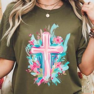 May include: A green t-shirt featuring a pink cross surrounded by blue and pink flowers. The cross is painted in a brushstroke style.