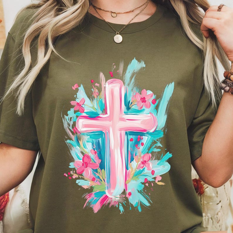 May include: A green t-shirt featuring a pink cross surrounded by blue and pink flowers. The cross is painted in a brushstroke style.