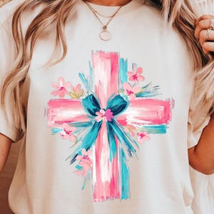 Puede incluir: Una camiseta blanca con un dise&ntilde;o de cruz pintada en rosa y azul. La cruz est&aacute; rodeada de flores rosas y un lazo azul.