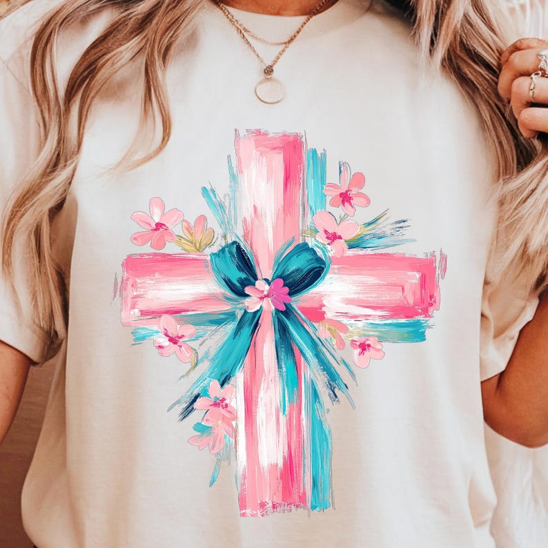 Puede incluir: Una camiseta blanca con un dise&ntilde;o de cruz pintada en rosa y azul. La cruz est&aacute; rodeada de flores rosas y un lazo azul.