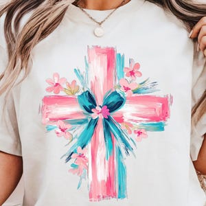 Puede incluir: Una camiseta blanca con un dise&ntilde;o de cruz pintada en rosa y azul. La cruz est&aacute; rodeada de flores rosas y un lazo azul.