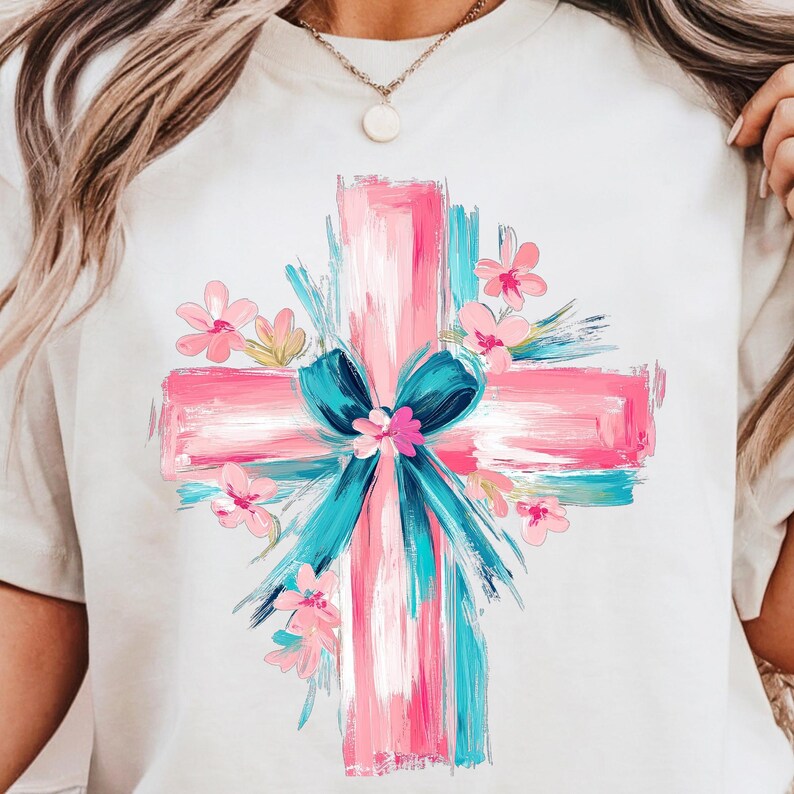 Puede incluir: Una camiseta blanca con un dise&ntilde;o de cruz pintada en rosa y azul. La cruz est&aacute; rodeada de flores rosas y un lazo azul.
