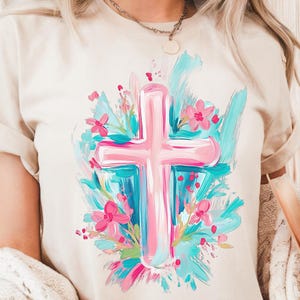 May include: A white t-shirt featuring a pink cross surrounded by blue and green brushstrokes and pink flowers.