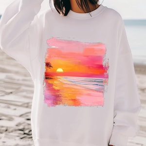 May include: White sweatshirt with a colorful sunset over the ocean graphic print. The graphic features a palm tree, a bright orange sun, and pink and orange clouds. The ocean is a light blue with white waves.