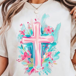 May include: A white t-shirt featuring a pink cross surrounded by blue and pink flowers. The cross is painted in a brushstroke style.