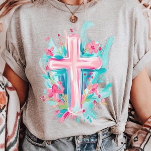 May include: A grey t-shirt featuring a pink cross surrounded by blue and pink flowers. The cross is painted in a brushstroke style.