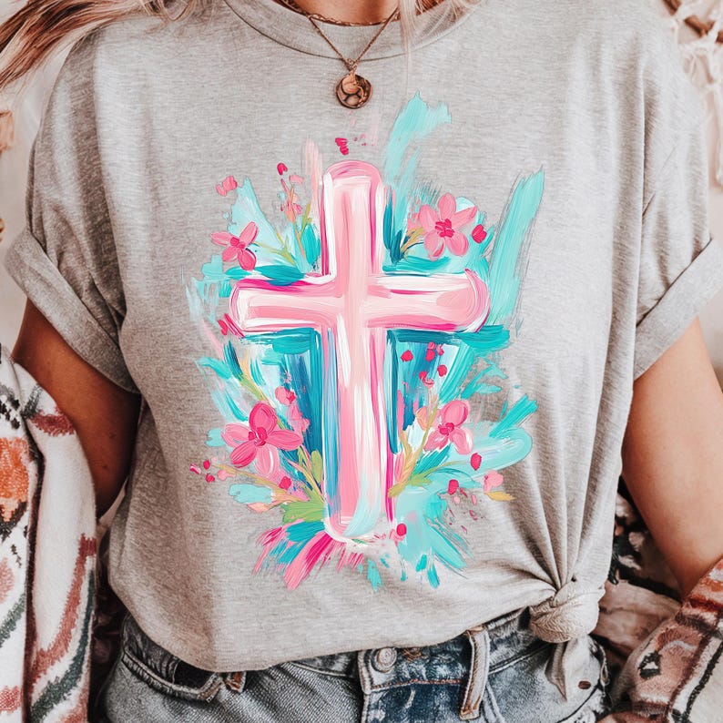 May include: A grey t-shirt featuring a pink cross surrounded by blue and pink flowers. The cross is painted in a brushstroke style.
