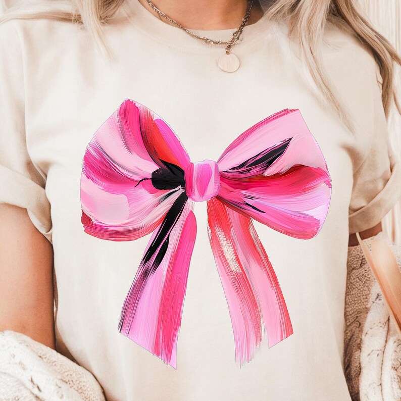 Puede incluir: Una camiseta blanca con un gran lazo rosa y negro pintado en la parte delantera. El lazo est&aacute; hecho de gruesas pinceladas de pintura rosa y negra.