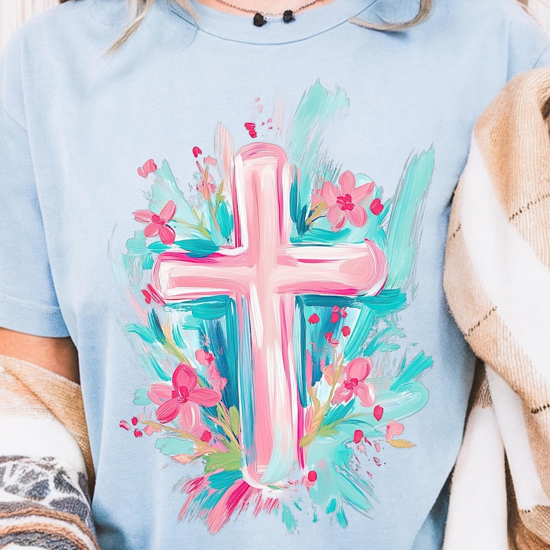 May include: A light blue t-shirt featuring a pink cross surrounded by blue and pink flowers. The cross and flowers are painted in a brushstroke style.