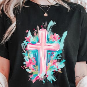 May include: A black t-shirt featuring a pink cross surrounded by blue and pink flowers. The cross is painted in a brushstroke style.