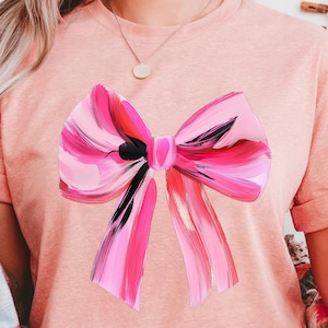 Puede incluir: Una camiseta rosa con un gran lazo rosa y negro pintado en la parte delantera.