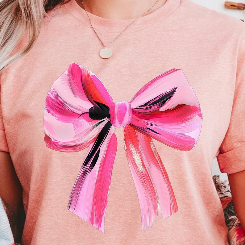 Puede incluir: Una camiseta rosa con un gran lazo rosa y negro pintado en la parte delantera.