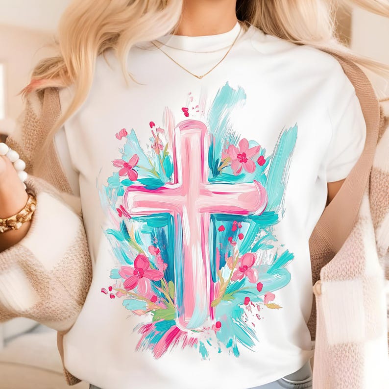 May include: White t-shirt featuring a pink cross surrounded by blue and pink flowers. The cross and flowers are painted in a brushstroke style.