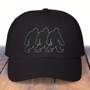 May include: Black baseball cap featuring an embroidered design of three Bigfoot silhouettes walking in a row. The cap has a curved brim and a structured fit, suitable for outdoor activities or casual wear.