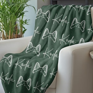 May include: A soft, green fleece throw blanket with a white repeating pattern of a dog's head and the name "Koda".